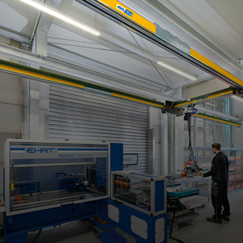 Enarmskranen EHB-X flyttar en last i en industrihall, medan en medarbetare arbetar vid en maskin i bakgrunden.